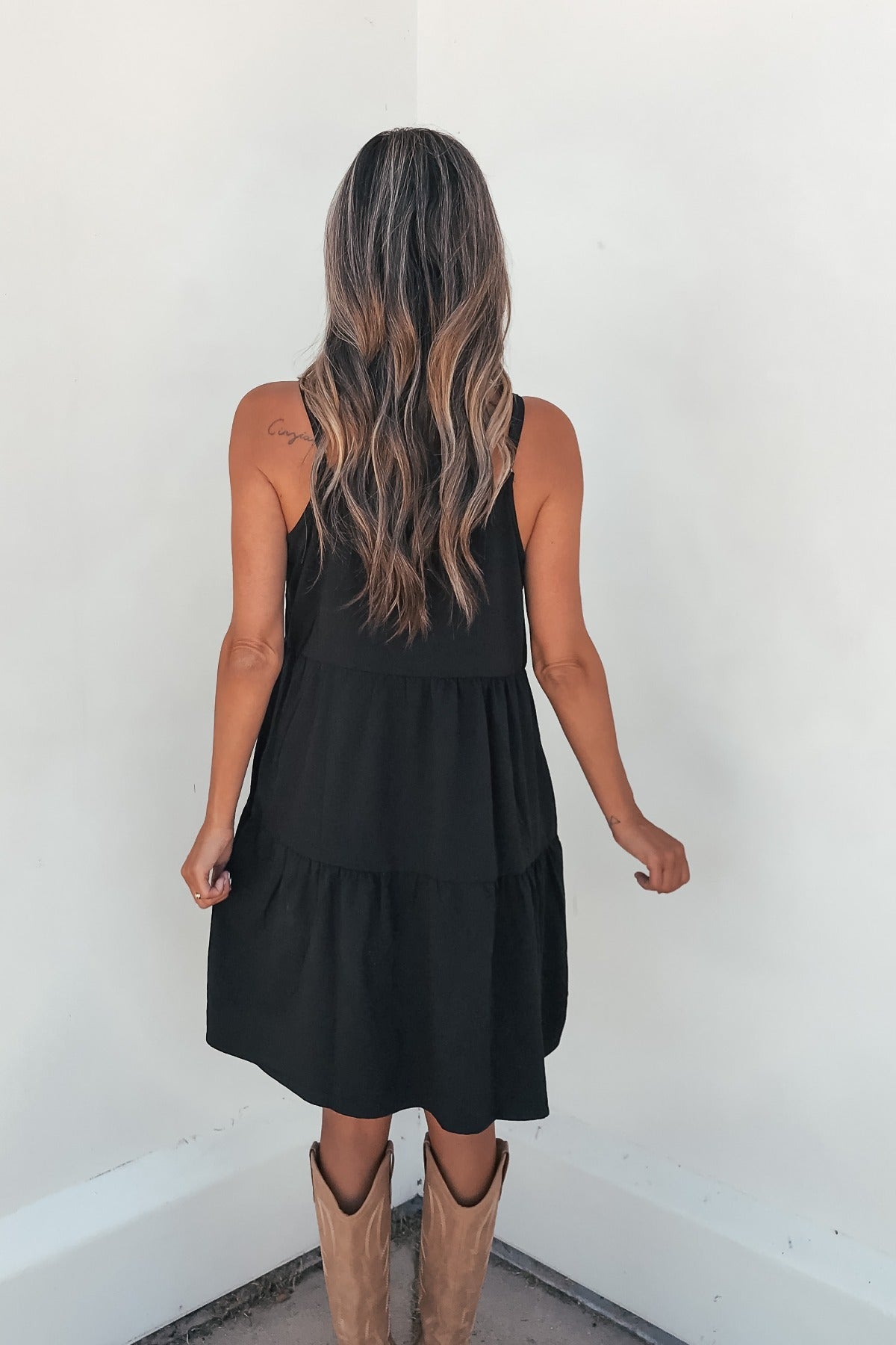 Woman with long wavy hair wears On Point Tiered Mini Dress - Black, styled with tan boots, standing against a white wall, back view.