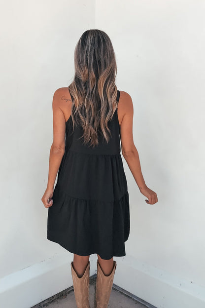 Woman with long wavy hair wears On Point Tiered Mini Dress - Black, styled with tan boots, standing against a white wall, back view.