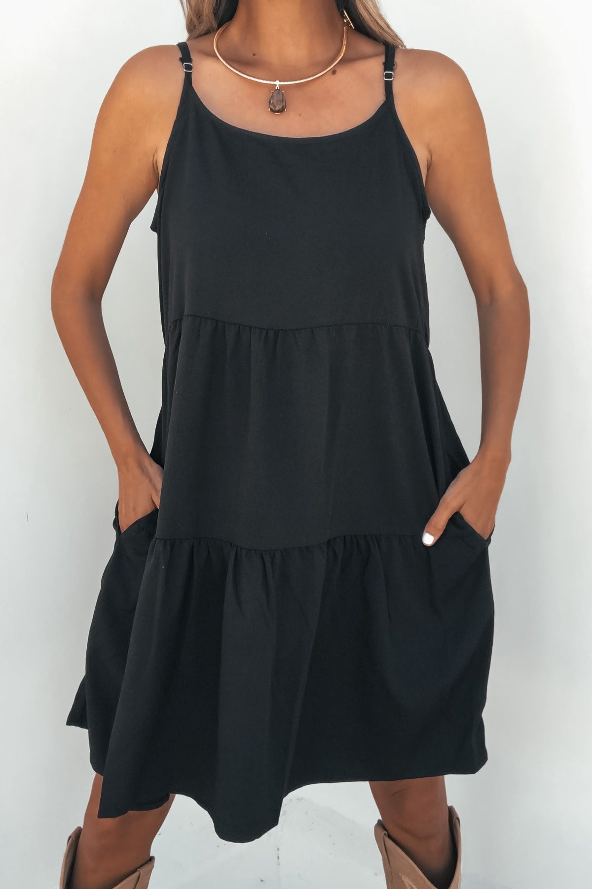 Model in On Point Tiered Mini Dress - Black, styled with a necklace and tan boots, poses against a white background.