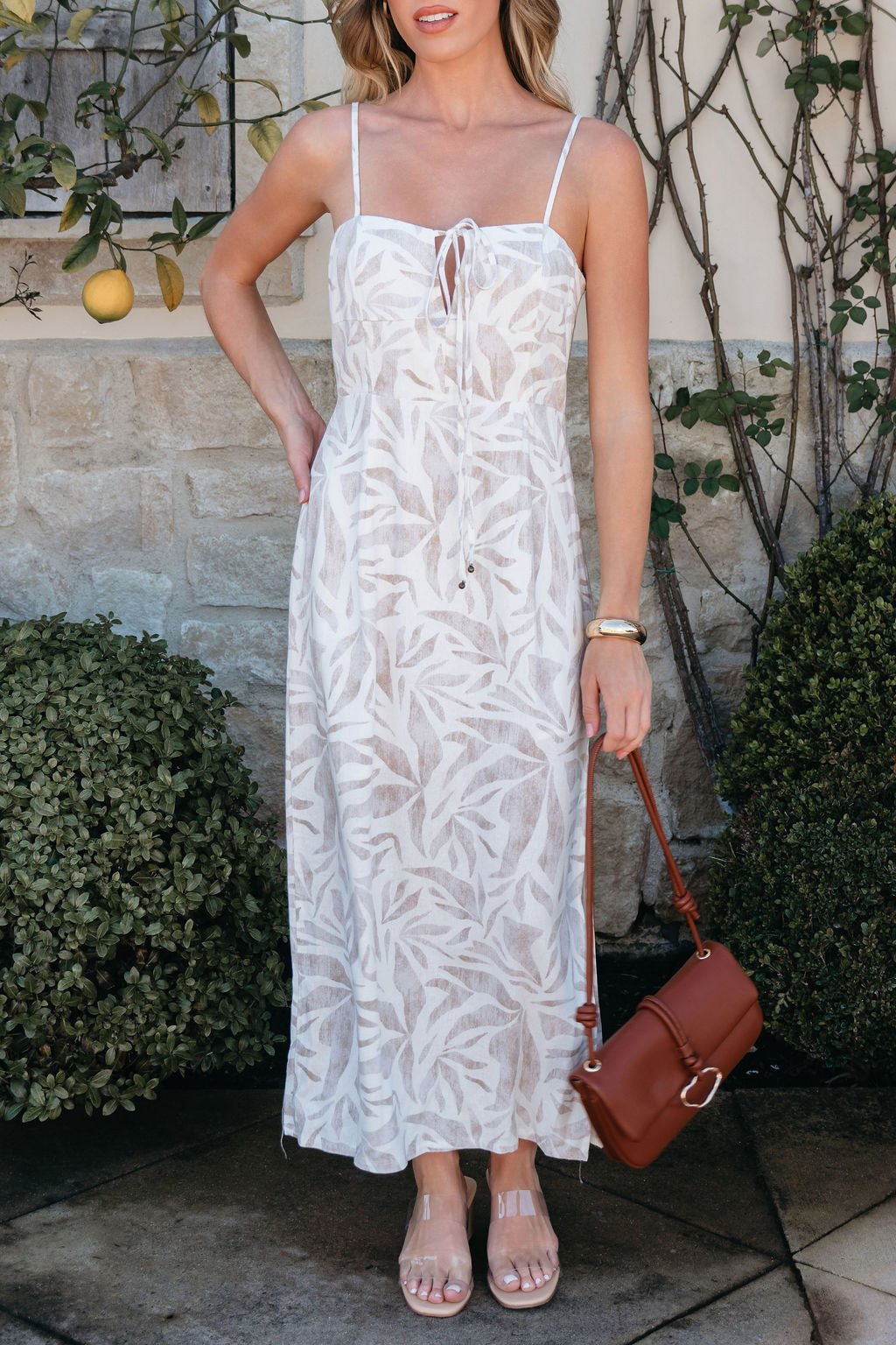 Woman in the Palm Vista White Maxi Dress (FINAL SALE) with clear sandals and brown handbag, outdoors by greenery and a stone wall.