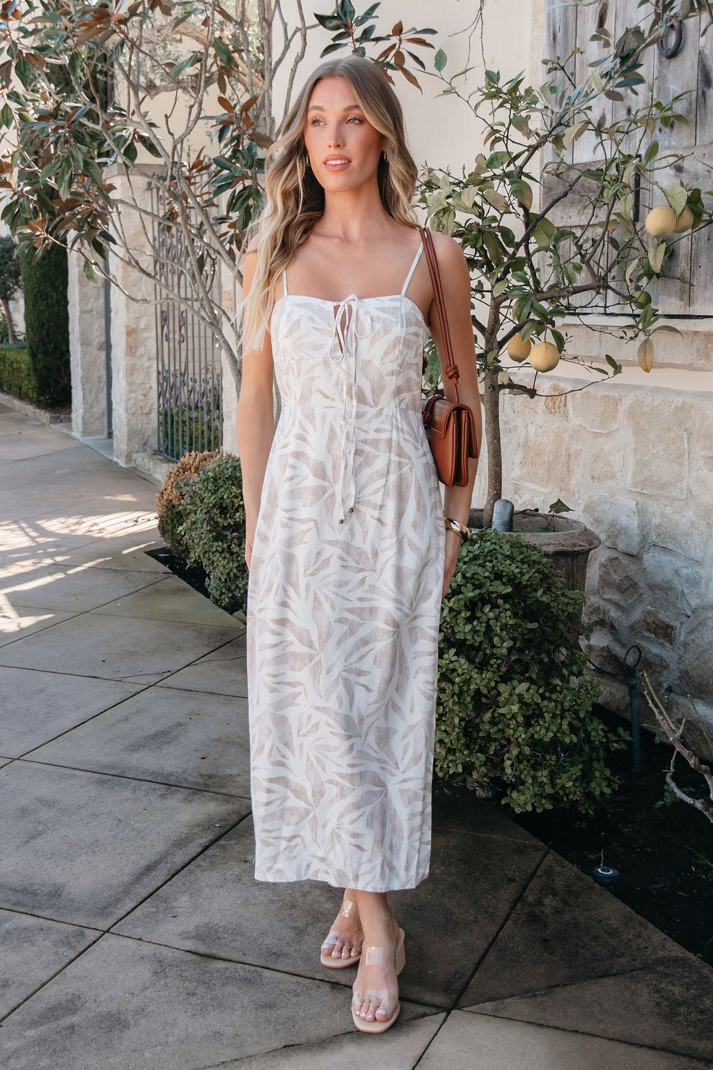 A woman stands outdoors with greenery, wearing the Palm Vista White Maxi Dress - FINAL SALE and holding a brown shoulder bag.