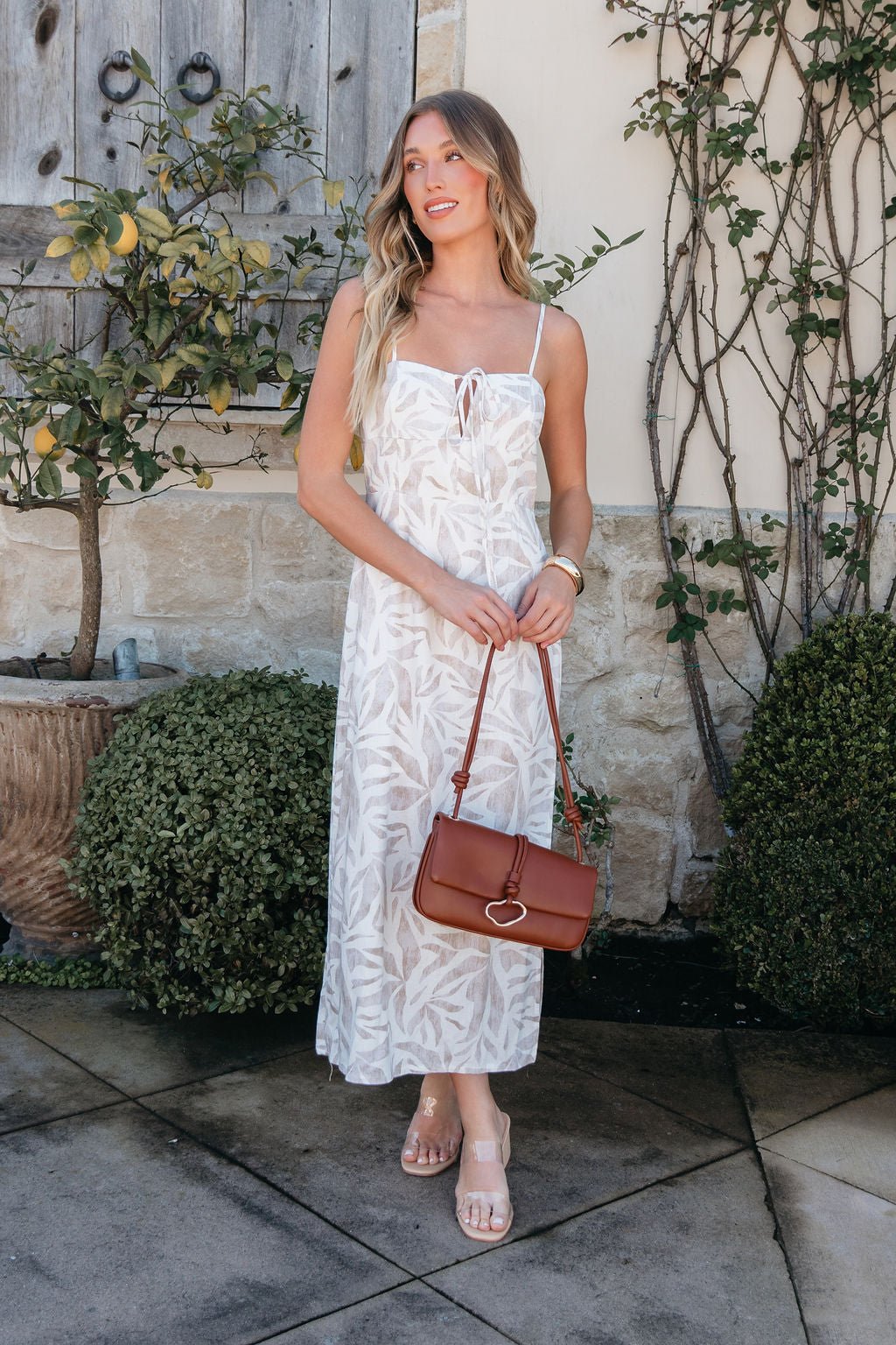 Woman wearing the Palm Vista White Maxi Dress, holding a brown purse by green shrubs and a stone wall with vines.