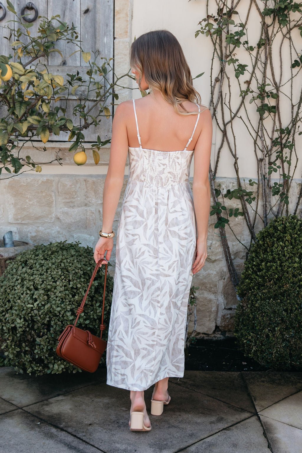Woman in the Palm Vista White Maxi Dress stands outdoors with sandals and a brown handbag, facing away from the camera.