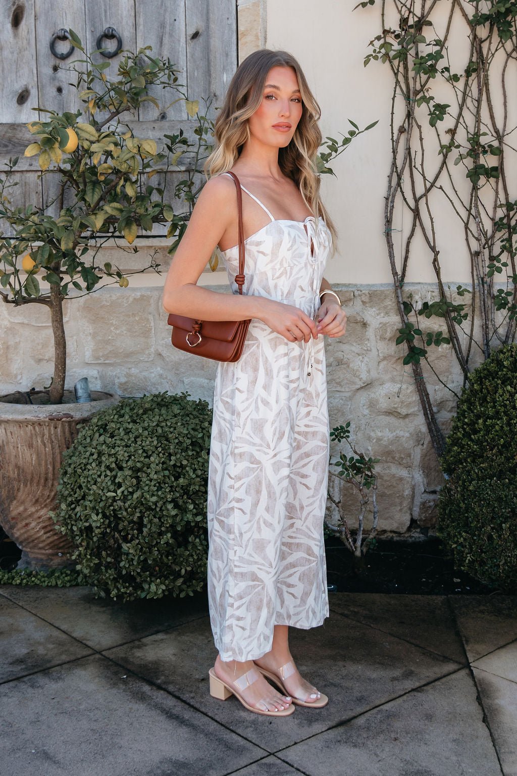 Woman in the Palm Vista White Maxi Dress stands outdoors by a stone wall and green plants.