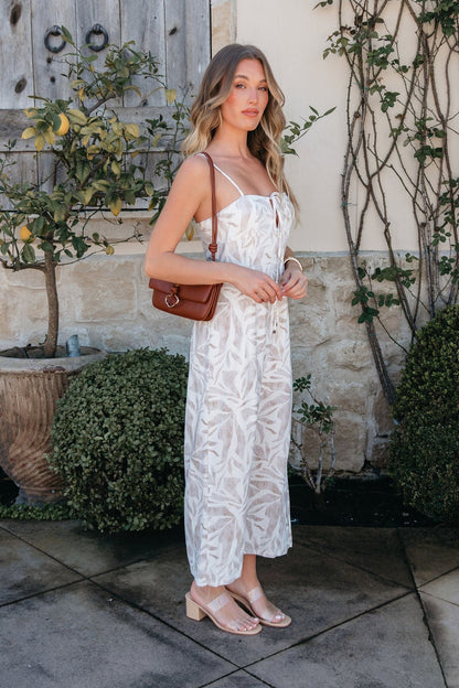 Woman in the Palm Vista White Maxi Dress stands outdoors by a stone wall and green plants.