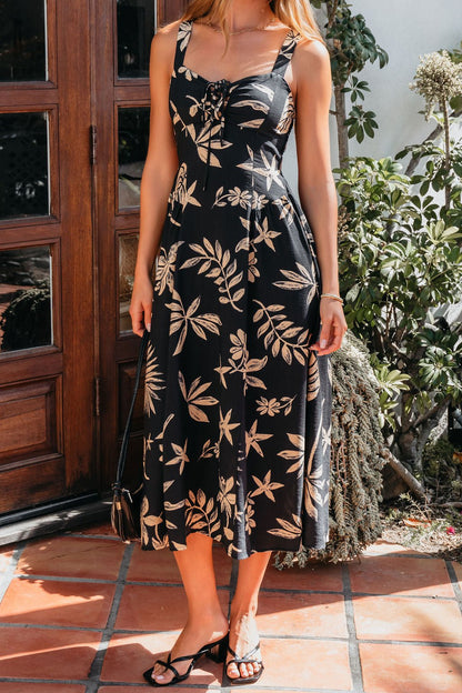 Woman in the Paloma Black Tropical Print Midi Dress, standing on a tiled patio near a wooden door and greenery.