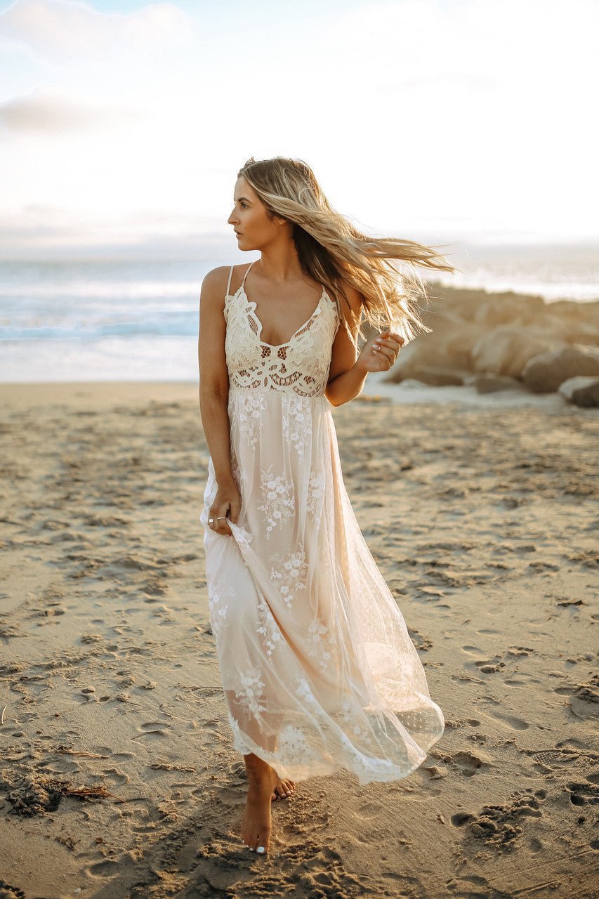 Woman in the Paradise Island Scalloped Vanilla Lace Maxi Dress walks barefoot on the beach at sunset, wind in her hair.