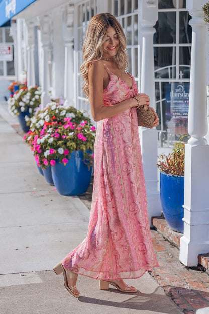 A woman smiles in the Pink Paisley Print Maxi Dress - FINAL SALE, walking past colorful potted flowers on the sidewalk.