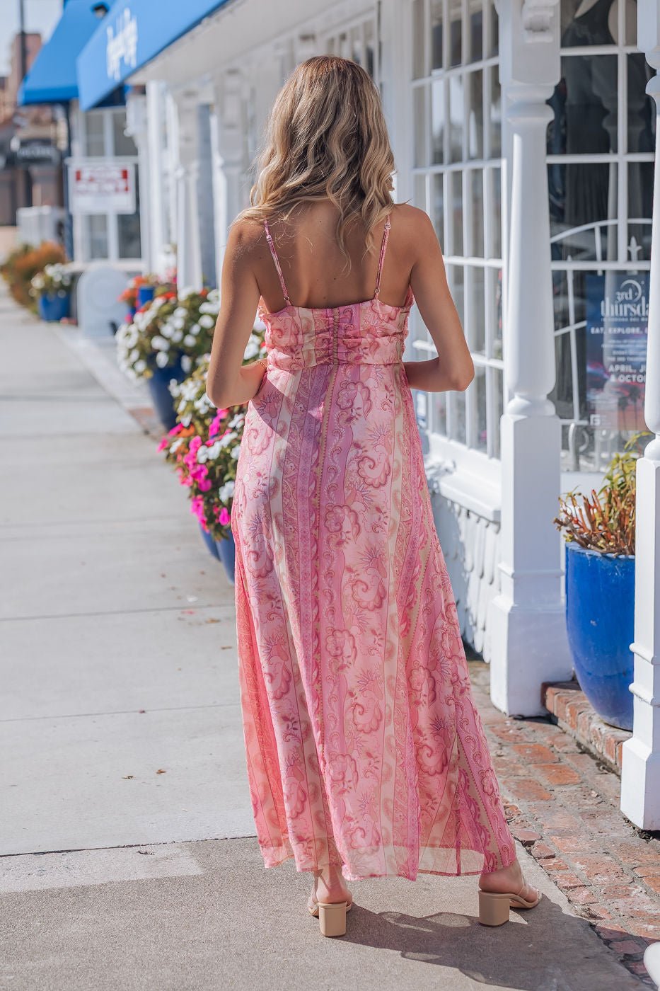 A woman strolls along sunny, flower-lined storefronts in the Pink Paisley Print Maxi Dress - FINAL SALE.