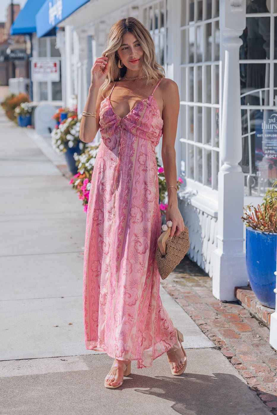 Woman in a Pink Paisley Print Maxi Dress - FINAL SALE, strolling in sandals with a woven clutch by white storefronts and flowers.