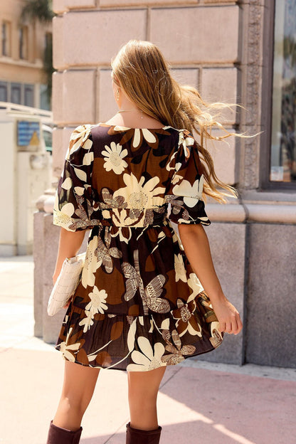 Woman wearing the Poppy Floral Mini Dress - Brown with puff sleeves and knee-high boots strolls city sidewalks, carrying a white purse.
