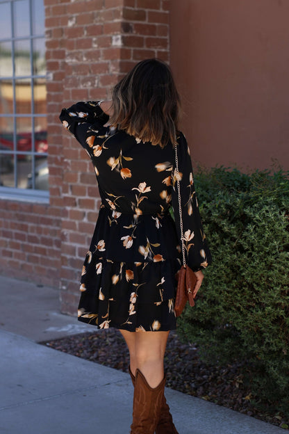 Woman in a Pumpkin Patch Black Floral Print Mini Dress and brown boots stands outside near a brick wall—a perfect fall look.