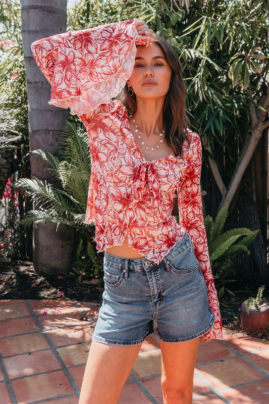 Woman poses outside in a Red Floral Print Bell Sleeve Mesh Top - FINAL SALE, styled with denim shorts amid lush green plants.