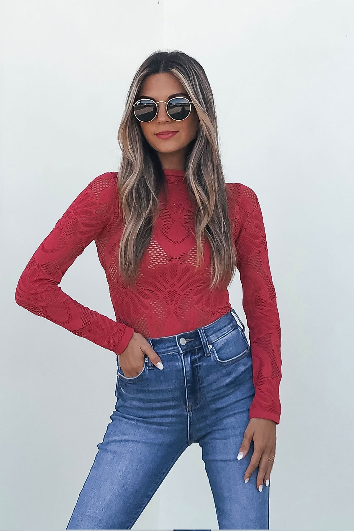 Woman in sunglasses models the Red Mock Neck Fishnet Top with high-waisted blue jeans on a simple light background.
