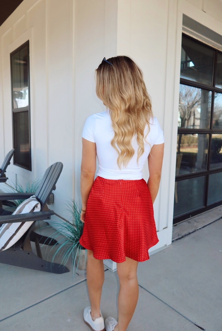 A woman in a white top and our Red Polka Dot Satin Mini Skirt - FINAL SALE stands outside by a white building with large windows.