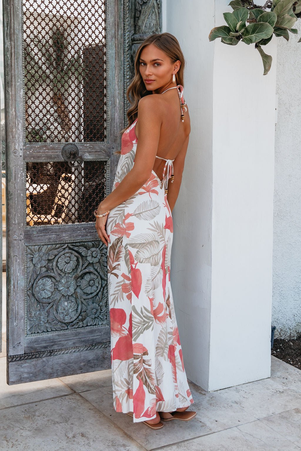 Woman in the Rosy Reef Escape Halter Maxi Dress poses by an ornate door, glancing over her shoulder at the camera.
