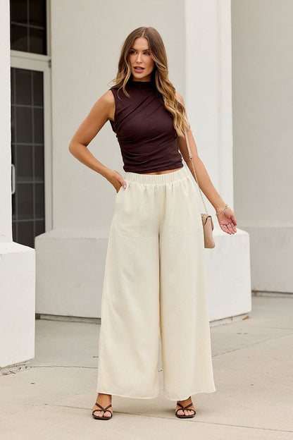 Woman in a Ruched Mock Neck Fitted Top - Brown, wide-leg cream pants, and beige purse stands outside a white building.