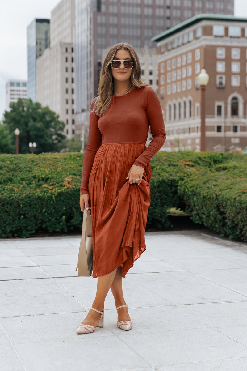 Woman in sunglasses and a Rust Contrast Pleated Midi Dress stands on a city sidewalk, holding a tan bag.