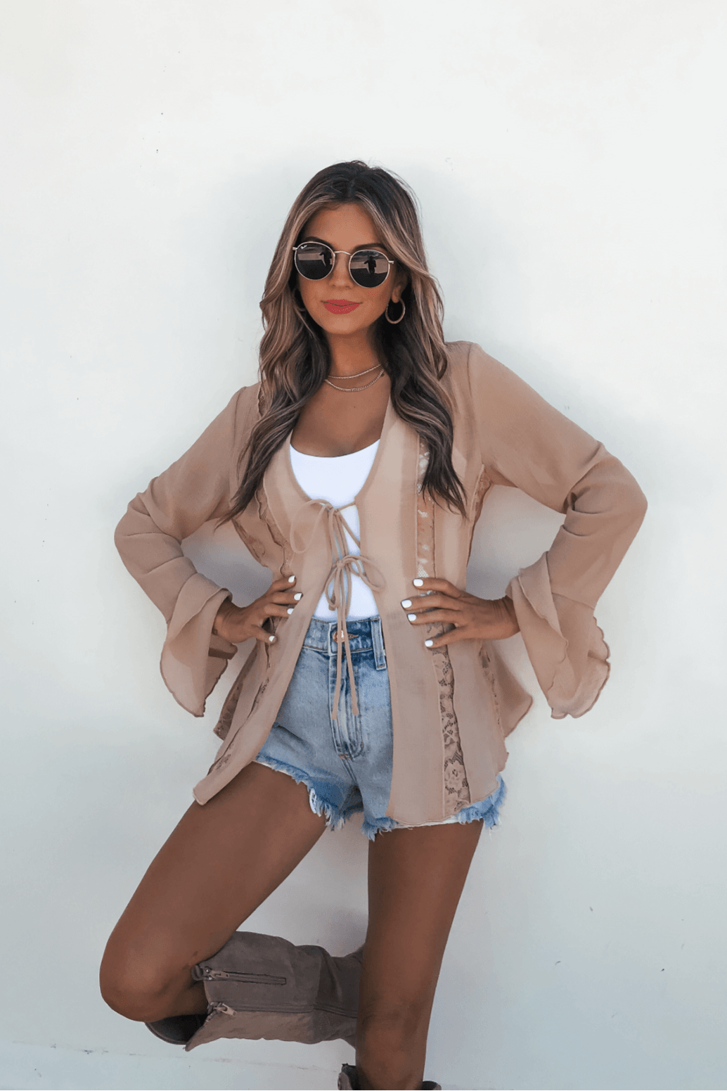 Woman poses in sunglasses, tan cardigan, Sand Bell Sleeve Sheer Lace Top, and denim shorts against a white wall with hands on hips.