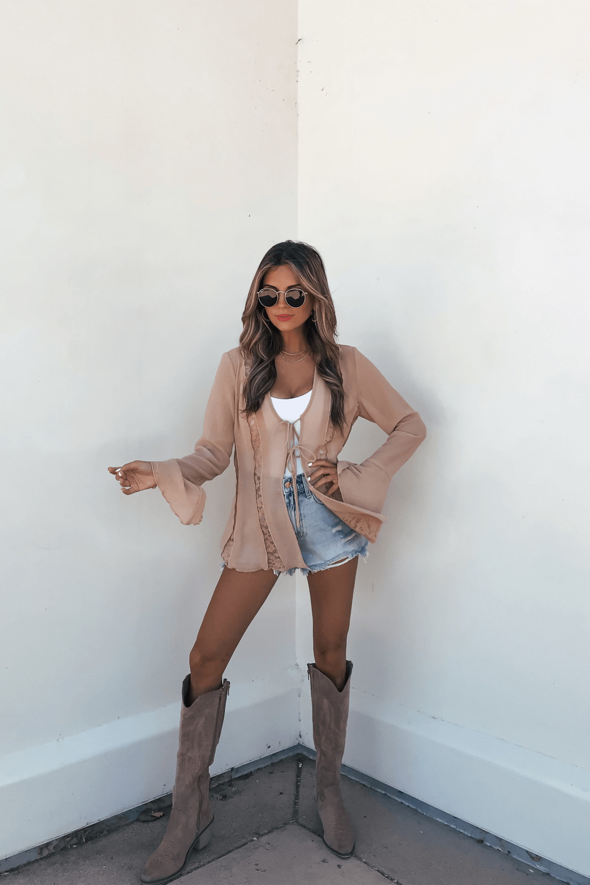 Woman in sunglasses wears the Sand Bell Sleeve Sheer Lace Top, denim shorts, and tan boots against a white wall.