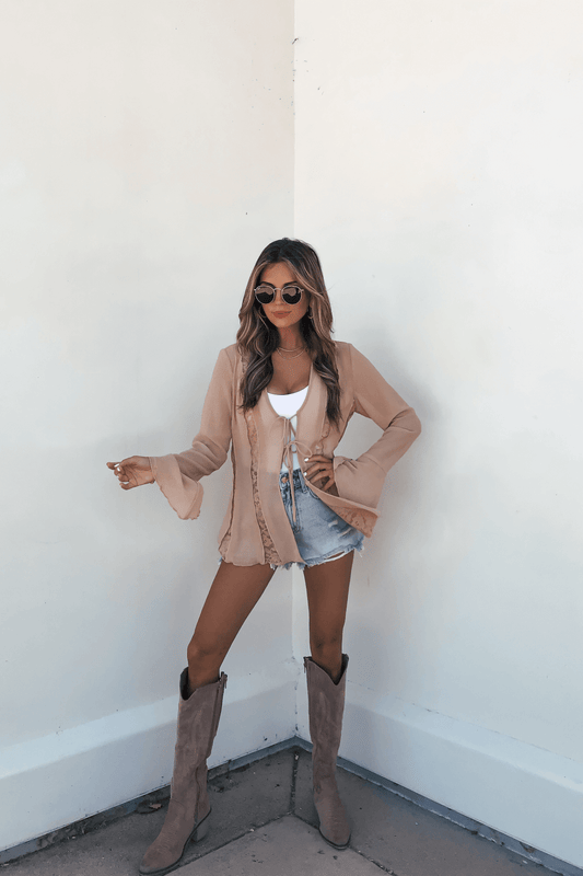 Woman in sunglasses wears the Sand Bell Sleeve Sheer Lace Top, denim shorts, and tan boots against a white wall.