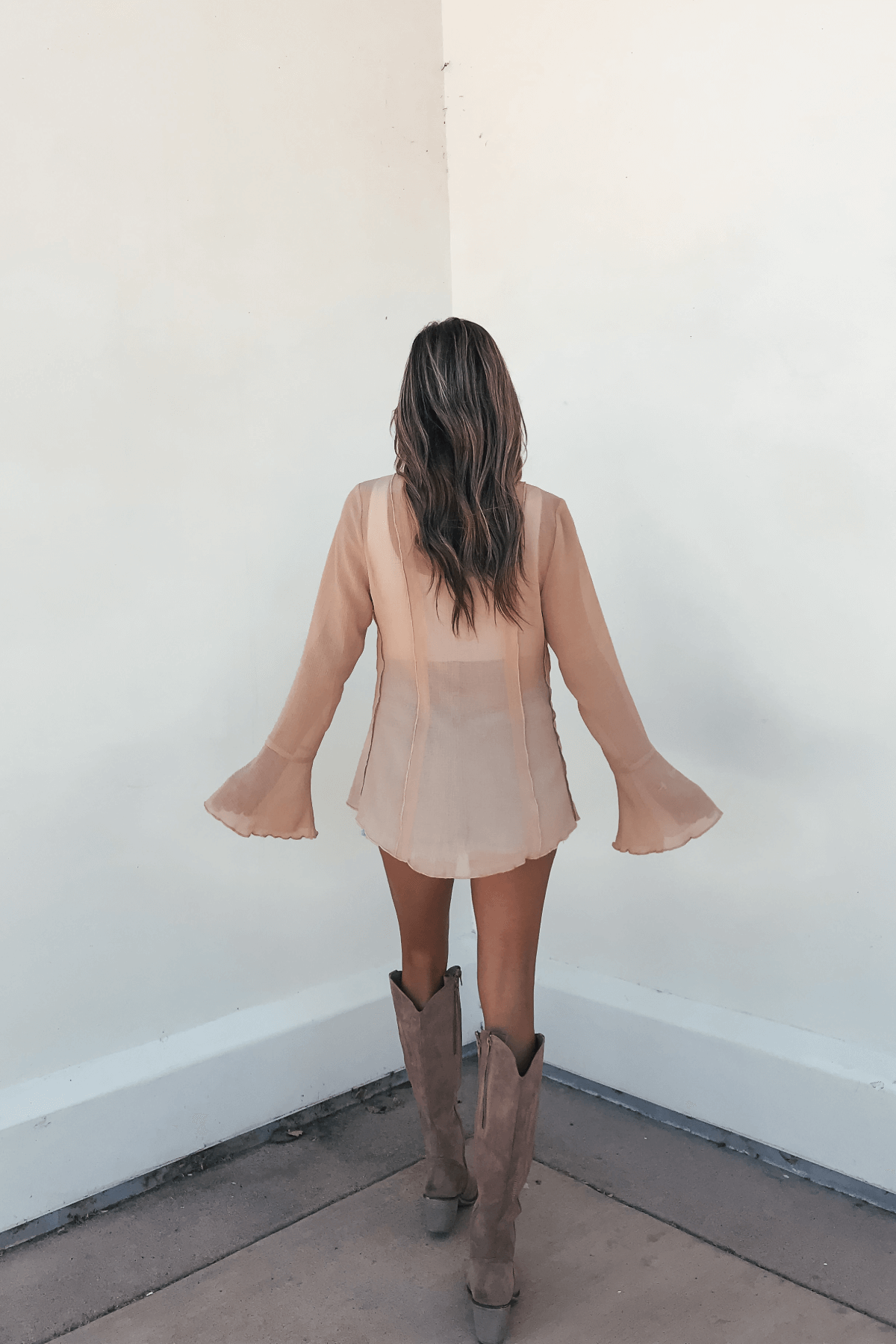 A woman with wavy hair wears the Sand Bell Sleeve Sheer Lace Top, standing on a concrete floor, facing a corner.