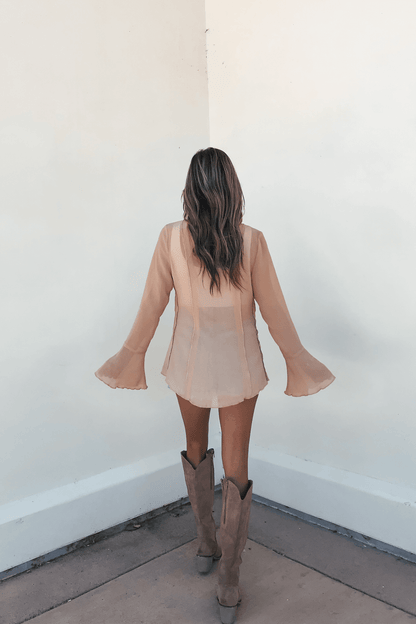 A woman with wavy hair wears the Sand Bell Sleeve Sheer Lace Top, standing on a concrete floor, facing a corner.