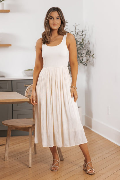 Woman in the Scoop Neck Pleated Skirt Midi Tank Dress stands indoors near a table and chair on a wooden floor.