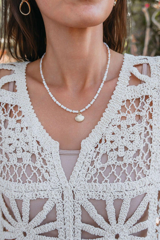 A woman wears a white crochet top and the Seabreeze Pearl Shell Pendant Necklace, featuring a beaded chain and gold shell charm.