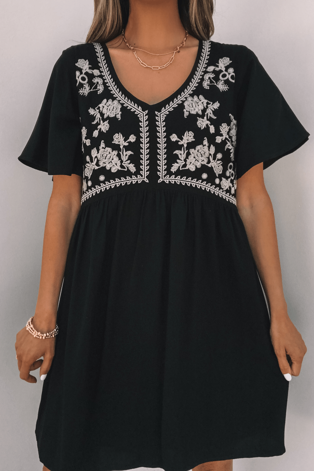 Woman in Seaside Embroidered Mini Dress - Black-FINAL SALE with white floral embroidery, short sleeves, and gold jewelry.