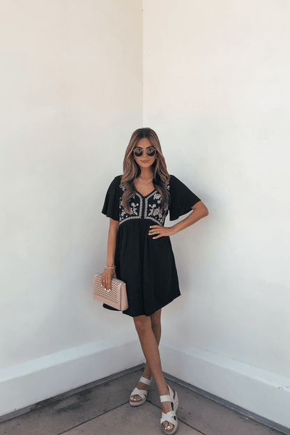 Woman in a Seaside Embroidered Mini Dress - Black-FINAL SALE, sunglasses, sandals, and quilted purse stands by a white wall.