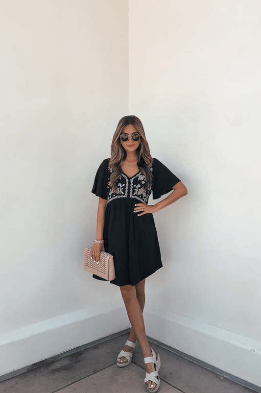 Woman in a Seaside Embroidered Mini Dress - Black-FINAL SALE, sunglasses, sandals, and quilted purse stands by a white wall.