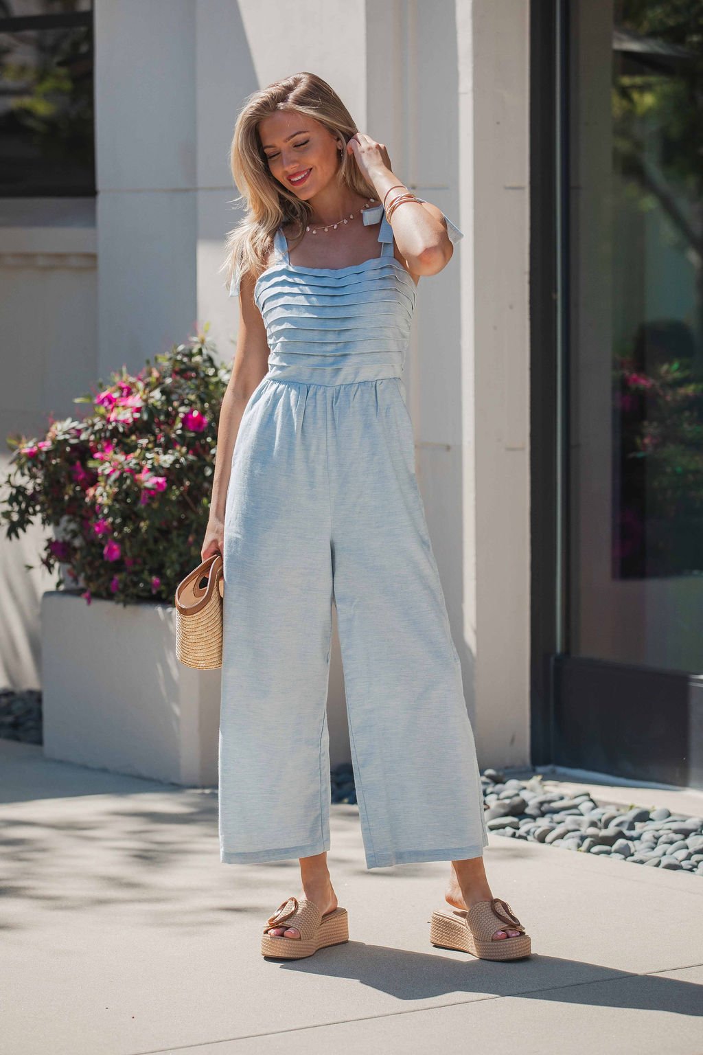 Woman in the Skylar Pleated Wide Leg Jumpsuit - Light Blue, with wedge sandals and a woven clutch, standing by flowers.