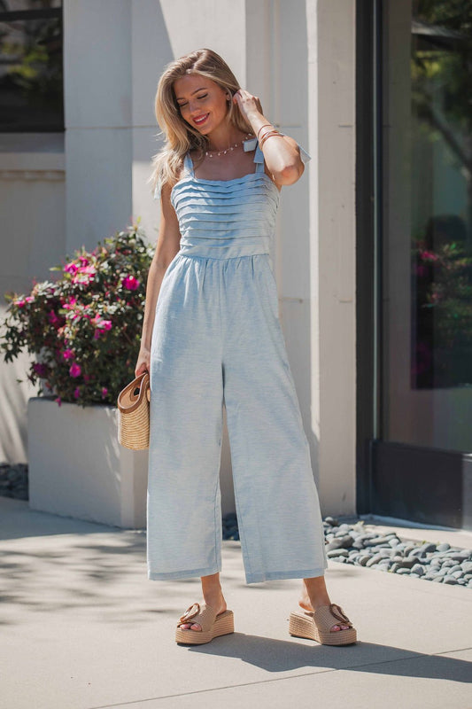 Woman in the Skylar Pleated Wide Leg Jumpsuit - Light Blue, with wedge sandals and a woven clutch, standing by flowers.