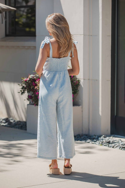 A woman in the Skylar Pleated Wide Leg Jumpsuit - Light Blue stands outside near a building with potted flowers.