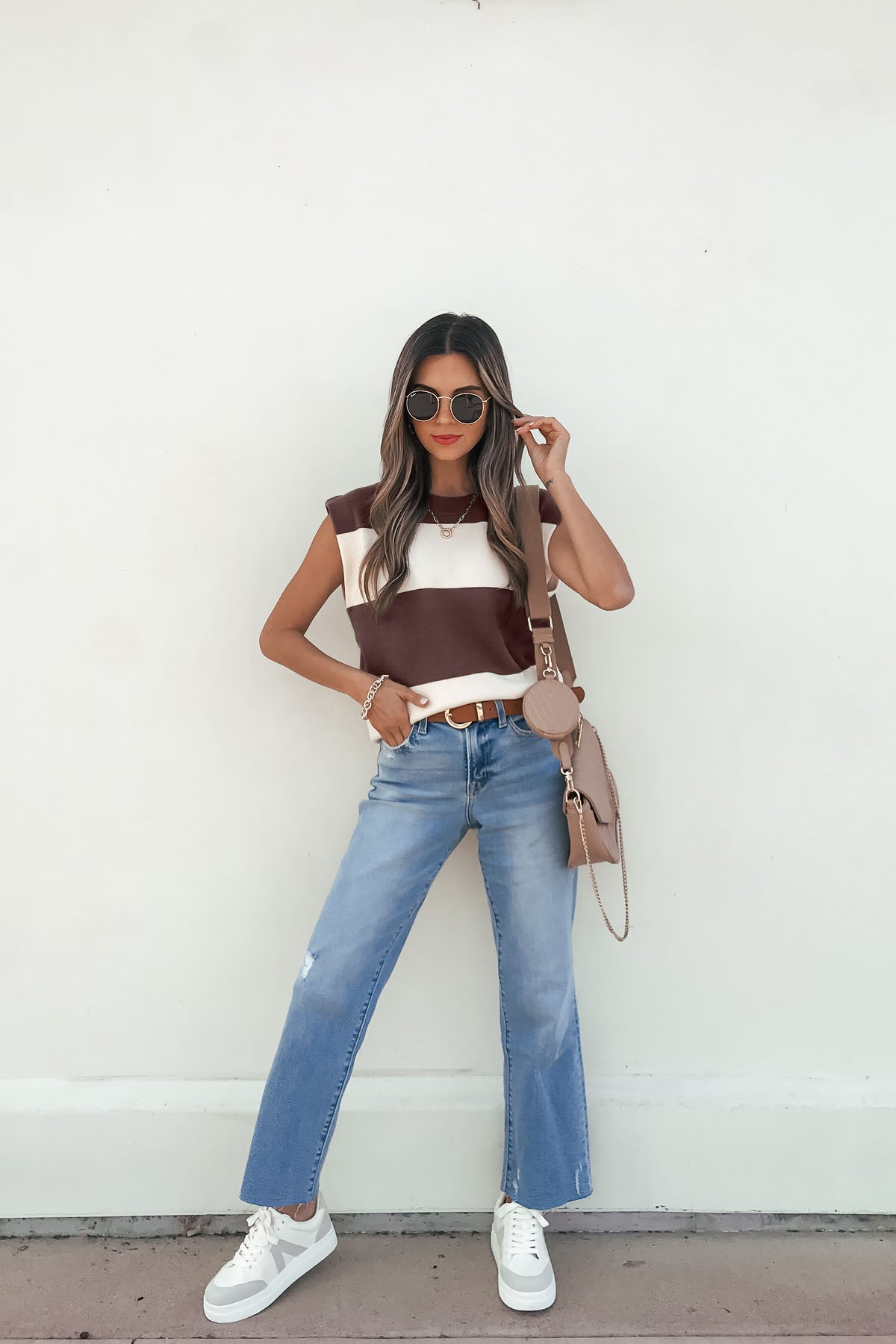 Woman in sunglasses wears a sleeveless brown and cream striped sweater, blue jeans, white sneakers, and a beige crossbody bag.