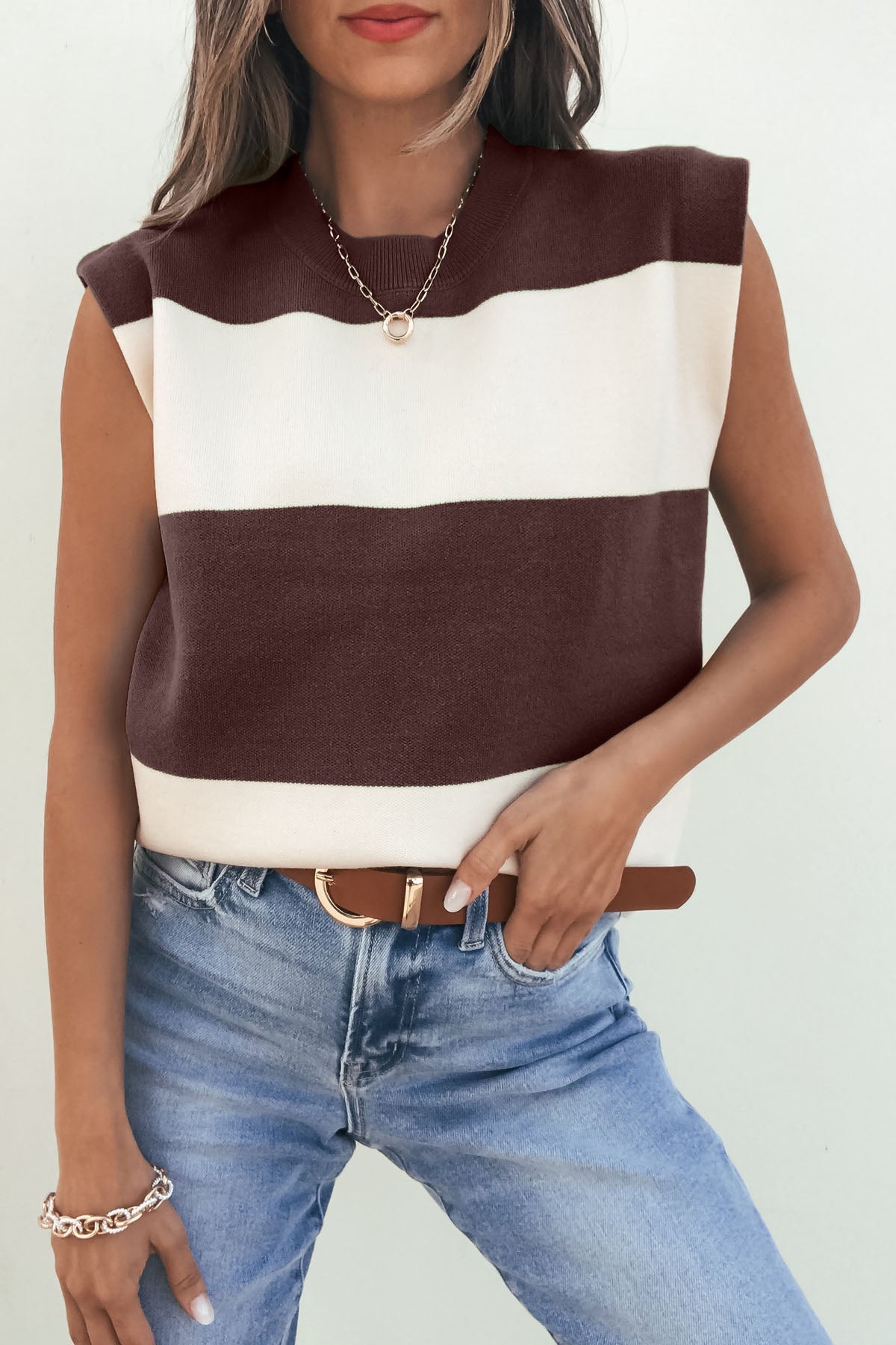Woman models a sleeveless brown and cream striped sweater with blue jeans, gold necklace, bracelet, and one hand in her pocket.