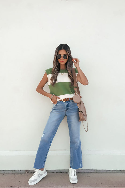 Woman in sunglasses, blue jeans, white sneakers, and a beige purse wears a Sleeveless Green and Cream Striped Sweater against a white wall.