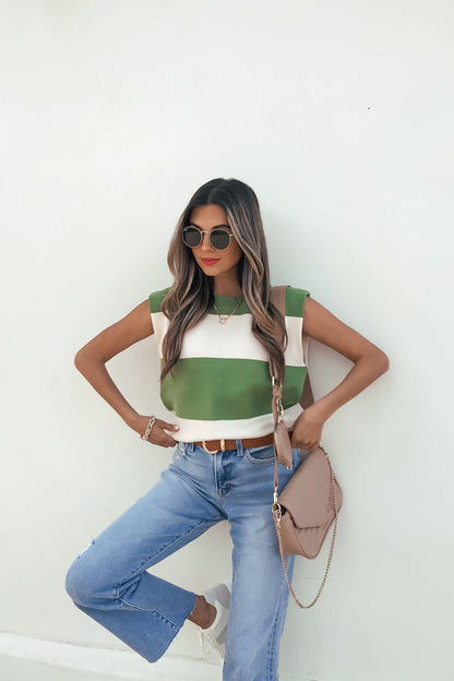 Woman in sunglasses and blue jeans models our Sleeveless Green and Cream Striped Sweater-FINAL SALE with a beige purse against a white wall.