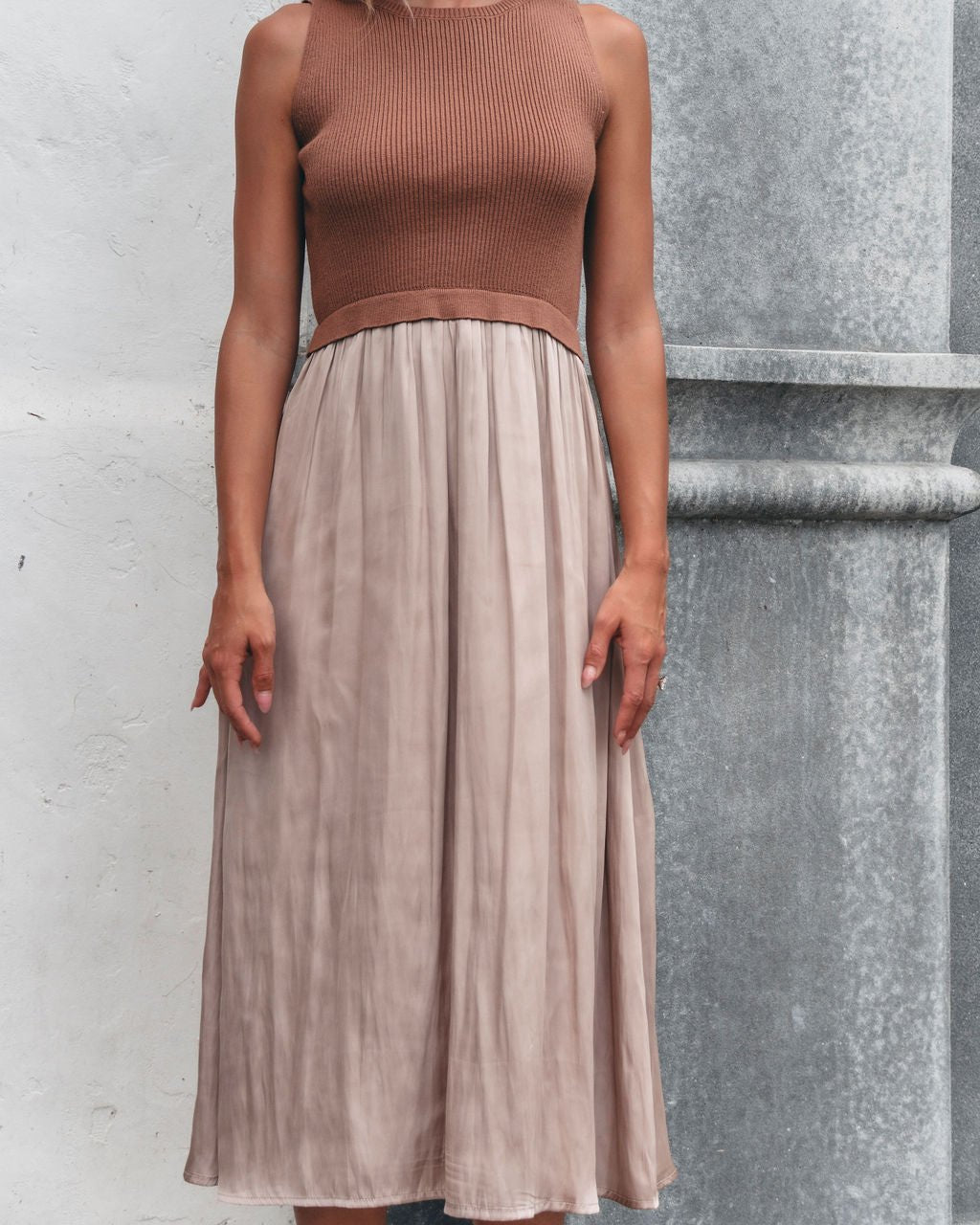 A woman in the Sleeveless Half Sweater and Satin Skirt Midi Dress - Mocha stands by a textured gray and white wall.