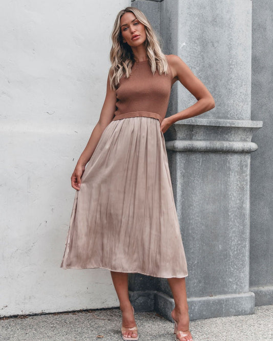 Woman in a Sleeveless Half Sweater and Satin Skirt Midi Dress - Mocha, stands against a wall, holding her skirt and looking at the camera.