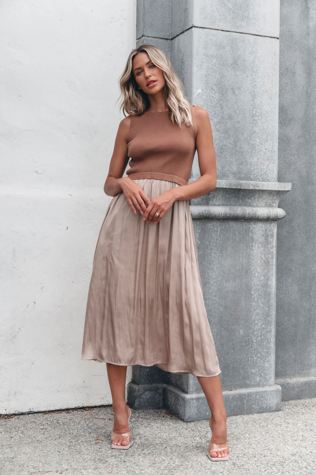 Woman wears the Sleeveless Half Sweater and Satin Skirt Midi Dress - Mocha against a gray stone pillar, paired with clear heels.