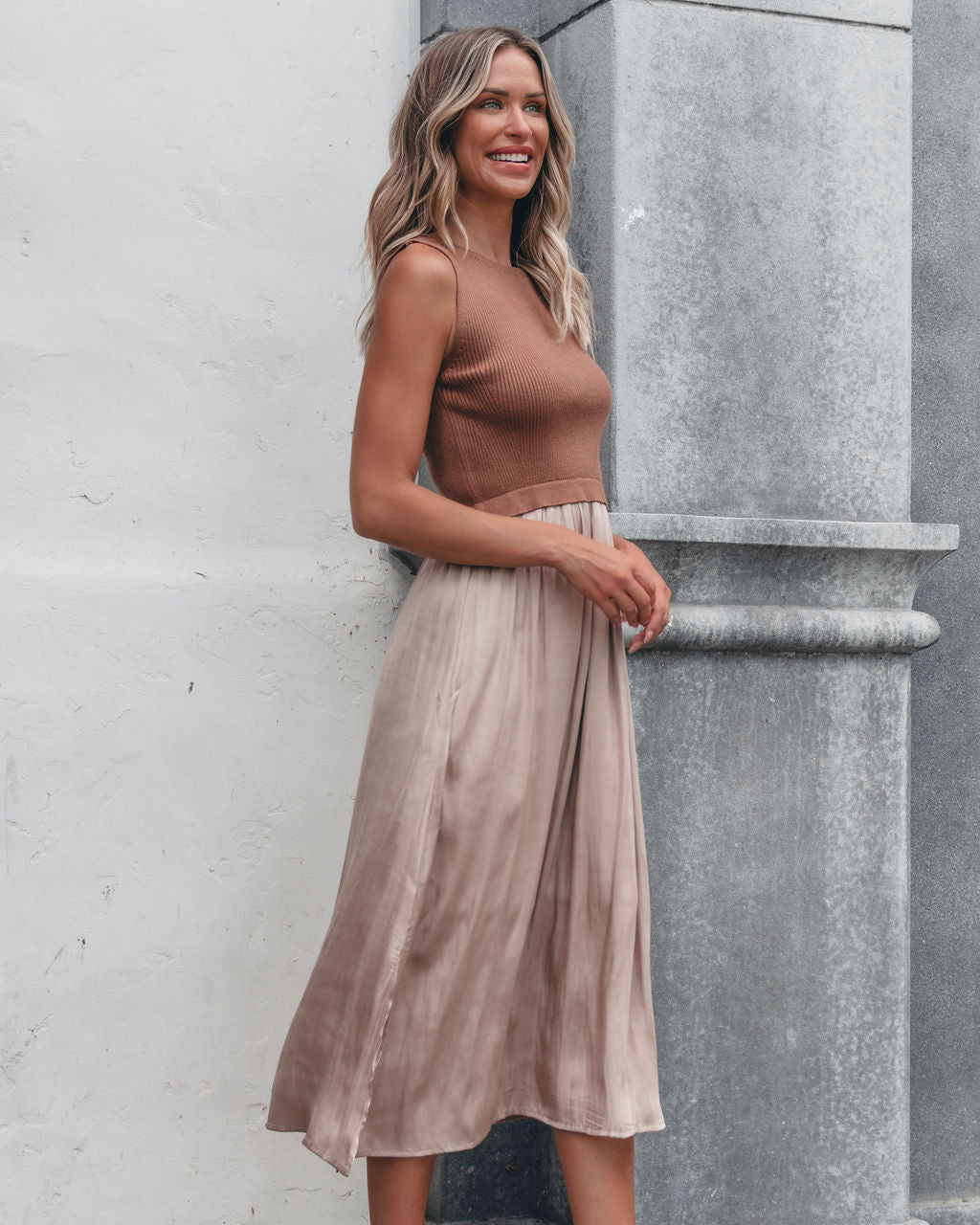Smiling woman in a Sleeveless Half Sweater and Satin Skirt Midi Dress in Mocha stands by a light gray wall.