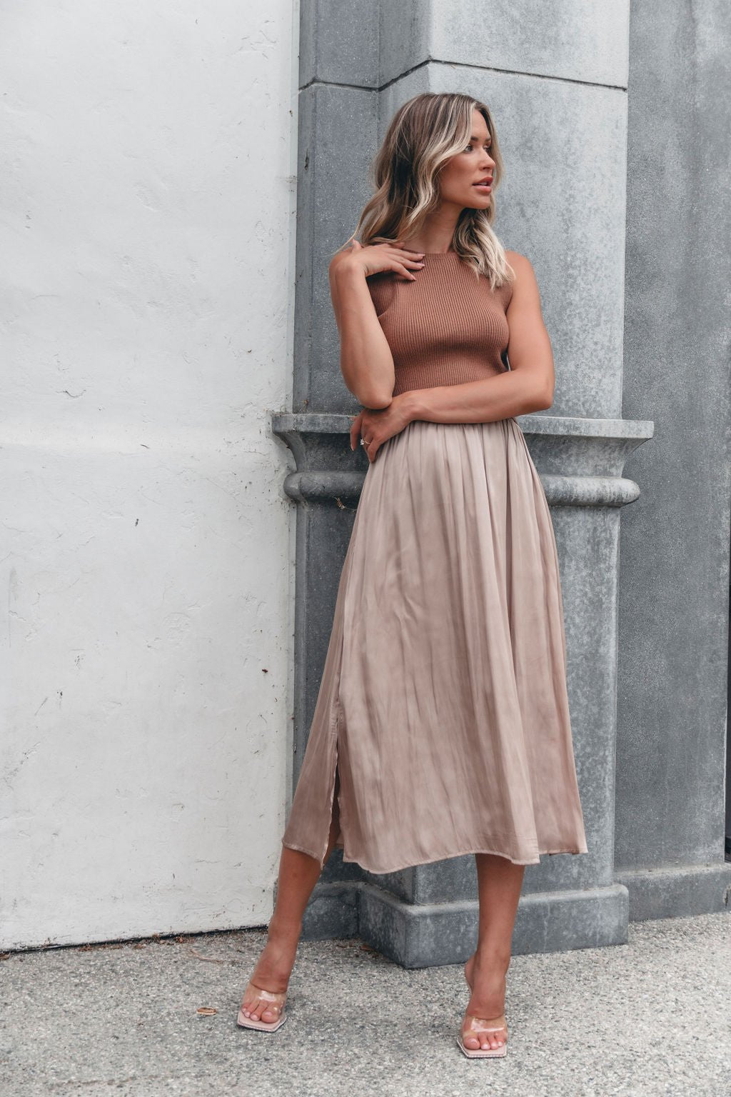Woman in the Sleeveless Half Sweater and Satin Skirt Midi Dress - Mocha stands by a stone pillar, gazing to the side.