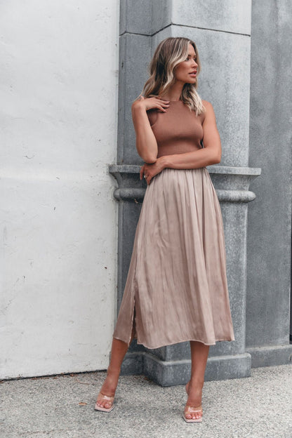 Woman in the Sleeveless Half Sweater and Satin Skirt Midi Dress - Mocha stands by a stone pillar, gazing to the side.