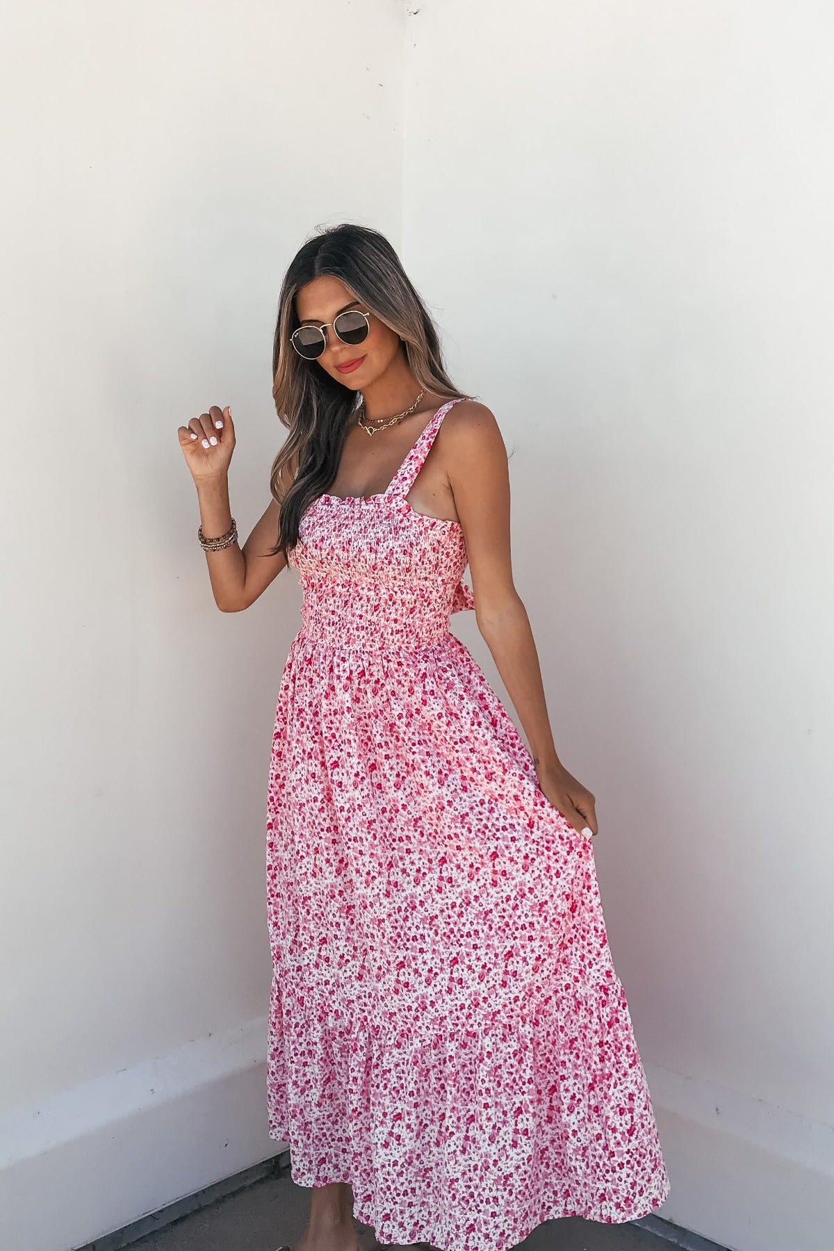 Woman in sunglasses wears the Smocked Bow Tie Print Midi Dress in pink, smiling as she stands against a white wall.