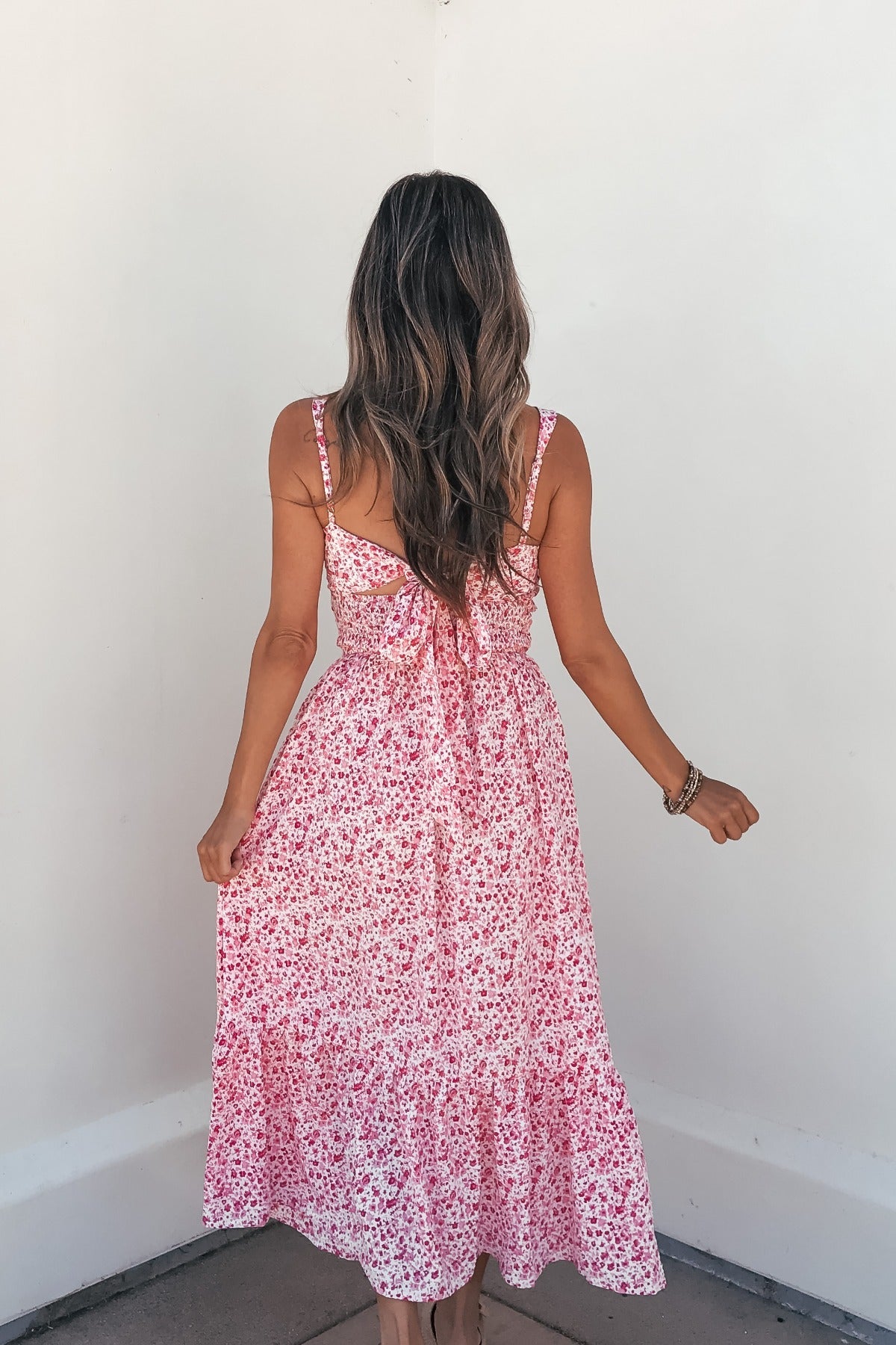 Woman in Smocked Bow Tie Print Midi Dress - Pink faces a white corner, holding her skirt.