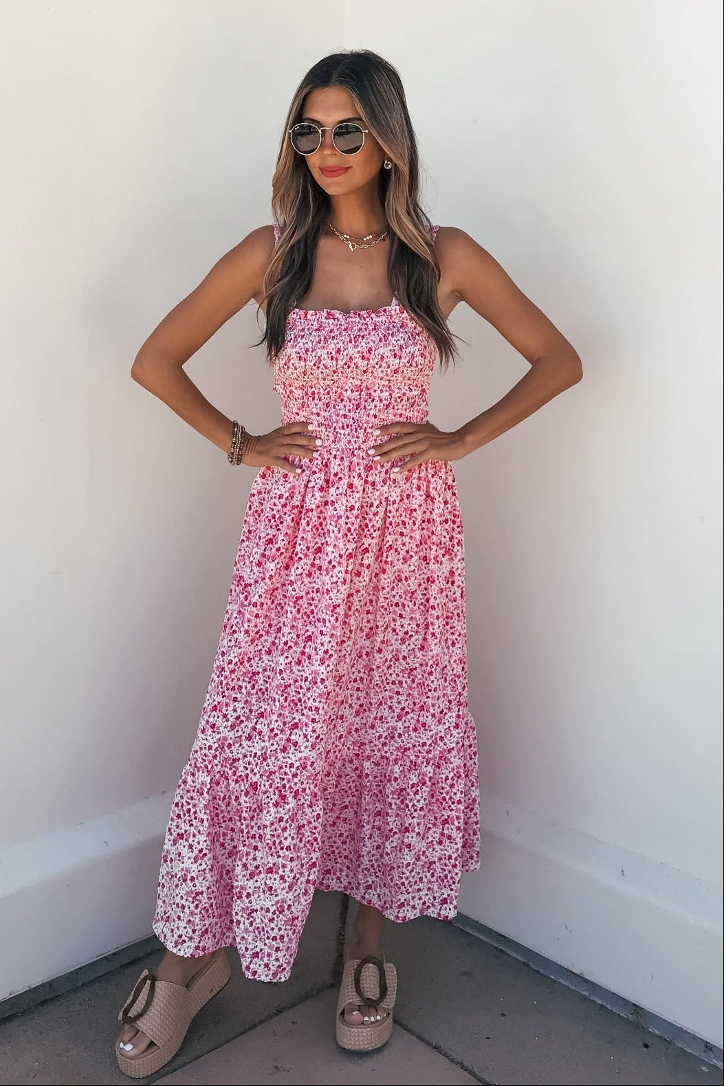 Woman wears Smocked Bow Tie Print Midi Dress - Pink, sunglasses, and wedges, posing against a white wall with hands on hips.