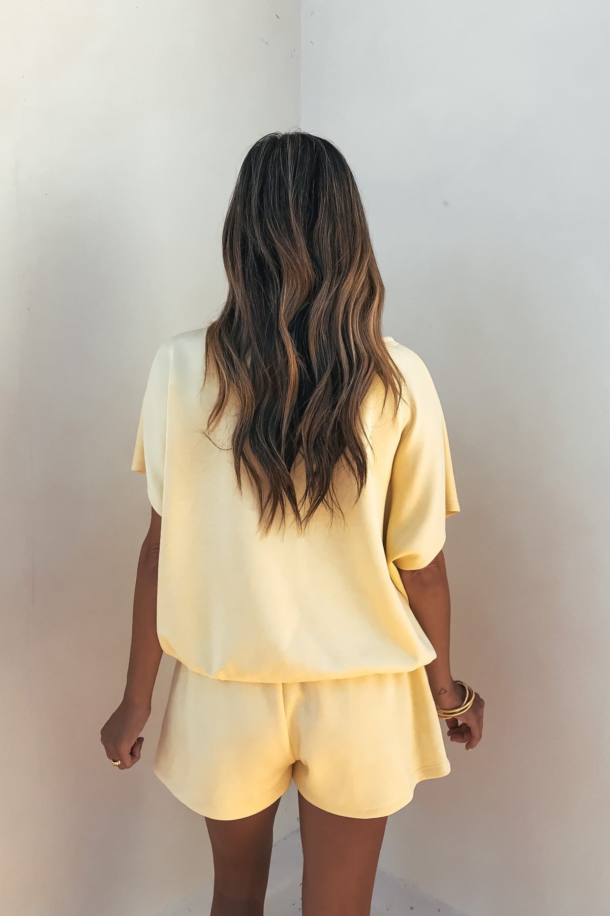 A woman wears Soft Cloud Scuba Lounge Shorts in yellow and a matching top, standing with her wavy hair down facing a white wall.