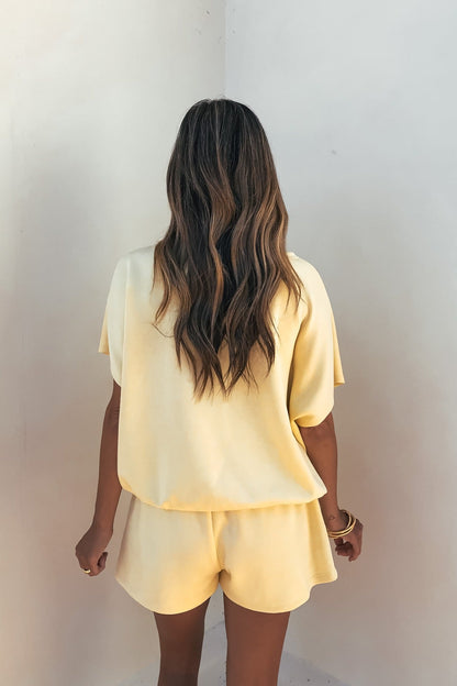 Woman with wavy brown hair wears the Soft Cloud Scuba Lounge Top in yellow, facing a white wall.